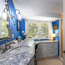Dated fixtures and kitschy wallpaper were unfortunate focal points prior to the cosmetic reimagining of this bathroom. The scalloped mirror provides an attractive centerpiece while the sconce lighting improves upon both visibility and aesthetics, while the updated cabinetry and countertops improve storage and surface area flexibility, retaining a convenient space for the vanity stool. A clean, modern blue now graces the walls as a timeless tile design refreshes the flooring. In combination with new Kohler hardware and sink, the bath now boasts an updated look on that classic bathroom feel.
